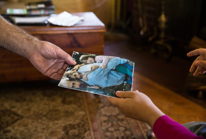 Photo: A close-up shot of Bernardo's hand holding an old picture of Bernardo, Gina, and her older sister. Gina's hand holds a corner of the photo.
