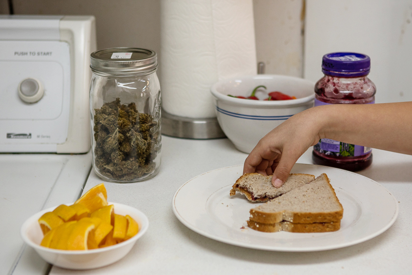 A woman holding a peanut butter and jelly sandwich. There are also orange slices and a jar filled with marijuana buds on the table.