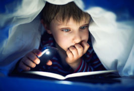 boy reading book under covers with flashlight
