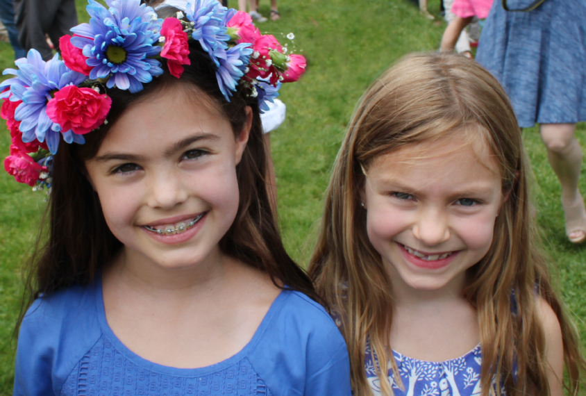 Photo: Two young girls smile at the camera. The girl on the left has long, dark brown hair and braces. She's wearing a blue shirt and a flower crown. The girl on the right has light brown hair and is wearing a floral blue and white shirt.