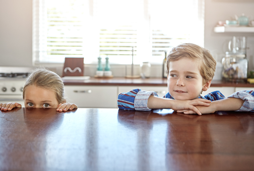 A young boy and a young girl hiding behind the kitchen table.
