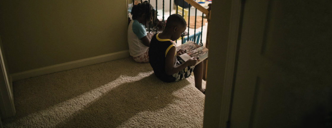 Photo: two children sit on the top step of a flight of carpeted stairs, reading size by side.
