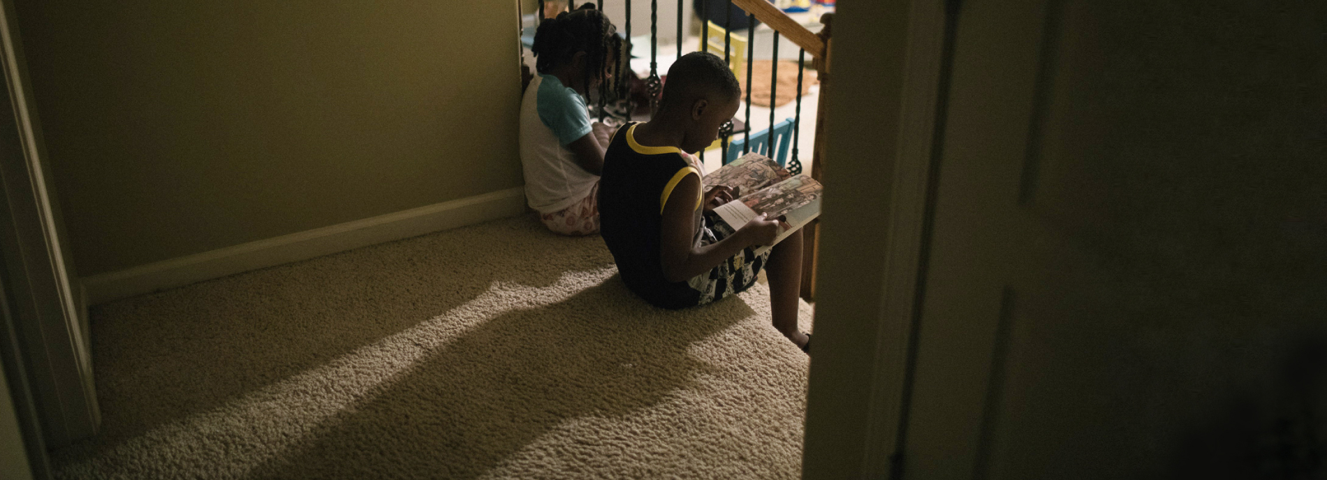 Photo: two children sit on the top step of a flight of carpeted stairs, reading size by side.