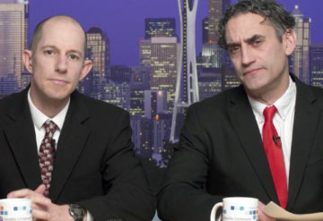 Two men dressed as news anchors look at the camera.
