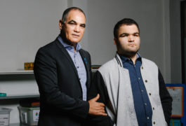 Photo: A father and son stand side by side, looking serious. The father is wearing a dark blue blazer over a grey sweater. The son is wearing a blue and grey jacket.