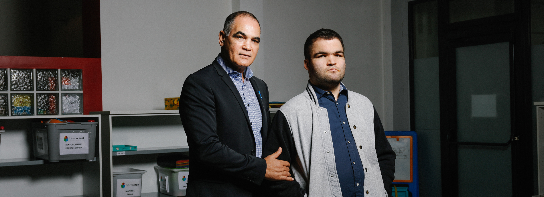 Photo: A father and son stand side by side, looking serious. The father is wearing a dark blue blazer over a grey sweater. The son is wearing a blue and grey jacket.