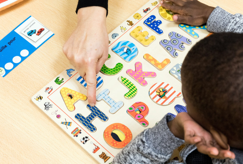 The photograph shows, from above, a child playing a game using cardboard letters fitting onto a board. An adult hand is in frame, pointing to the letter H.